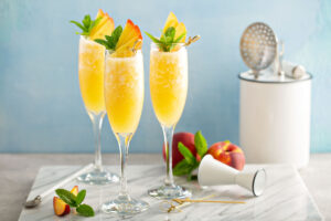Mimosa bar with three champagne glasses filled with fresh peach mimosas on a marble cutting board
