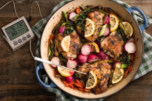 Digital thermometer stuck in chicken thigh in enameled cast iron dish on distressed wood table