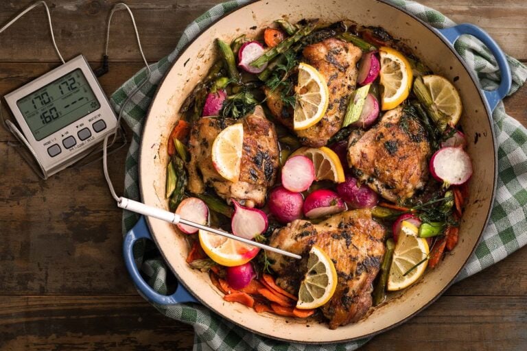 Digital thermometer stuck in chicken thigh in enameled cast iron dish on distressed wood table